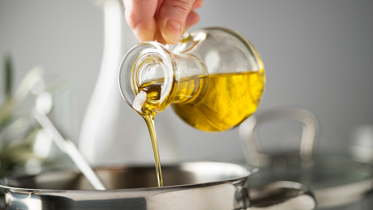 A hand pour out olive oil of a glass into a kitchen pot in the kitchen.